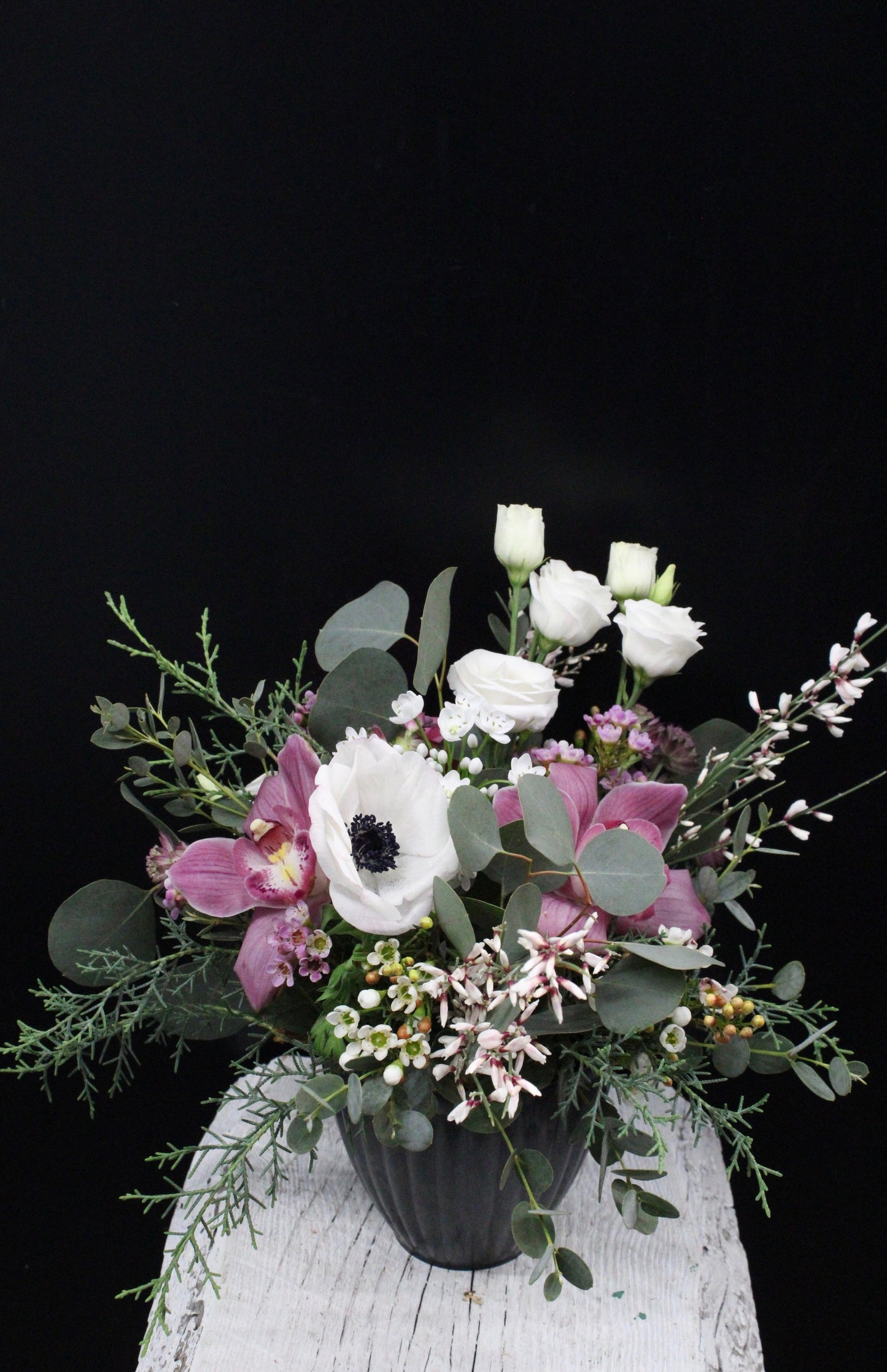 A floral arrangement featuring orchids, white anemone with black center, white roses, and greenery, presented in a container.