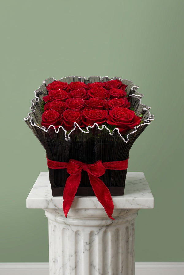 A square floral arrangement of 16 red roses in a black box, wrapped in black paper with a white beaded scalloped edge and tied with a red velvet ribbon bow, set on a wooden tabletop.
