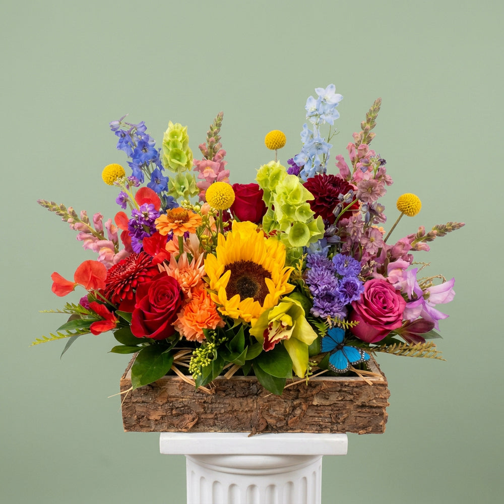 A vibrant, multicolor floral arrangement featuring a large yellow sunflower, red and purple roses, orange gerbera daisies, and green foliage, presented in a rustic wooden box on a counter inside a flower shop.