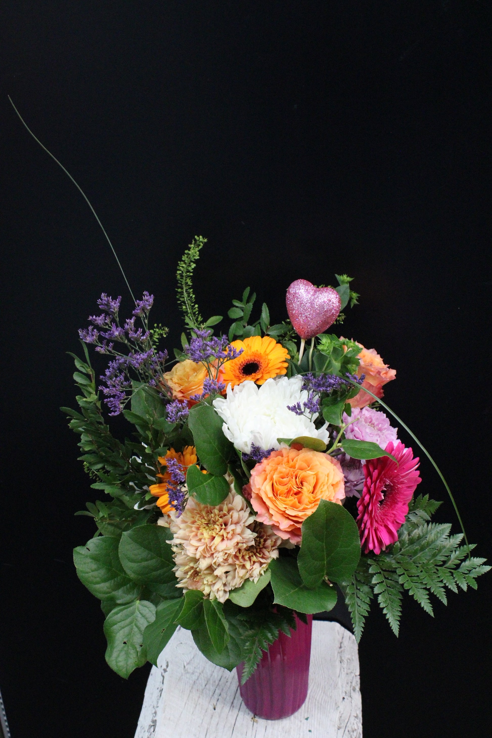 A vibrant bouquet of orange and pink gerbera daisies, with orange roses and purple accents, arranged in a vase with a decorative pink heart.