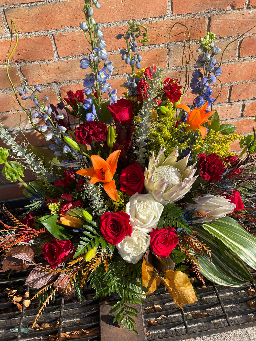 A vibrant floral arrangement featuring a variety of flowers including red roses, orange lilies, and white tulips, with greenery and decorative elements in between, displayed against a brick wall background.